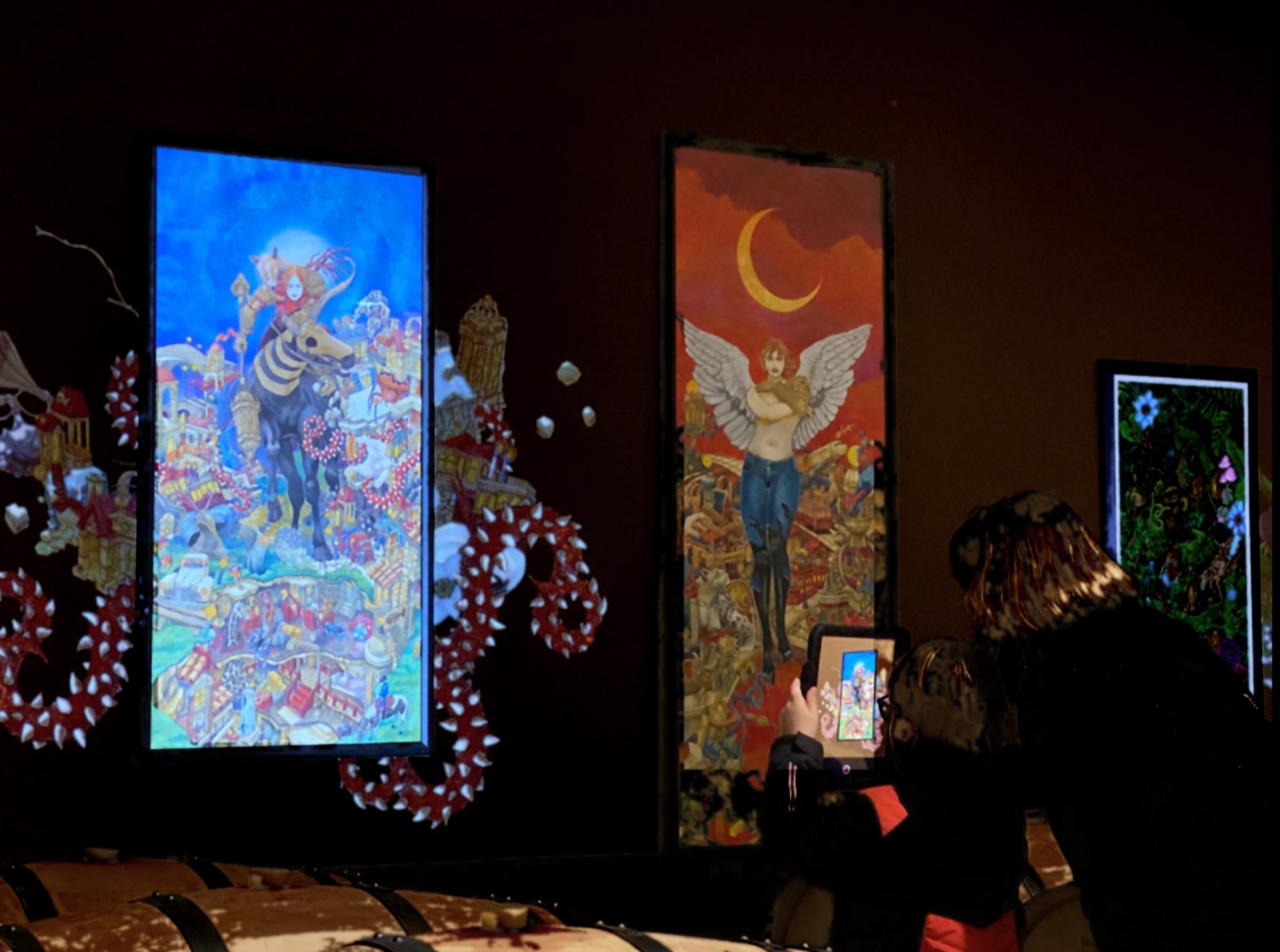 Vue de l'exposition en ambiance sombre : les illustrations de Grégory Dayon se prolongent au-delà de leurs cadres et une visiteuse découvre une œuvre en réalité augmentée sur tablette
